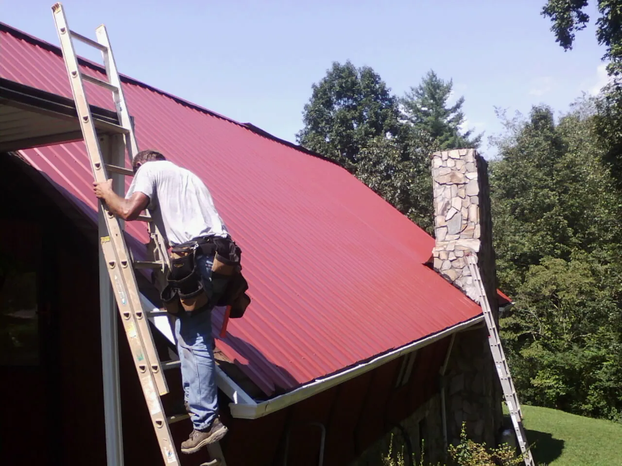 Roofer on a ladder beside a red metal roof and stone chimney — Soffit & Fascia Repair by Roofing Brothers in Orangeburg